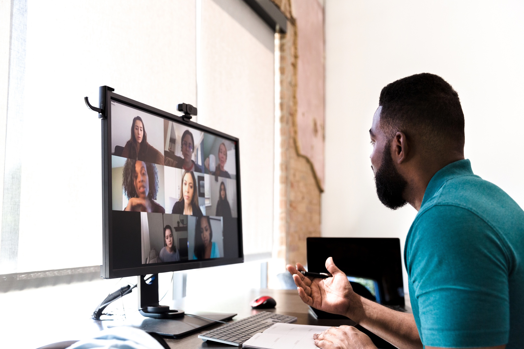 Photo of A mid adult man gestures while talking with a group of colleagues during a virtual staff meeting via video conference.